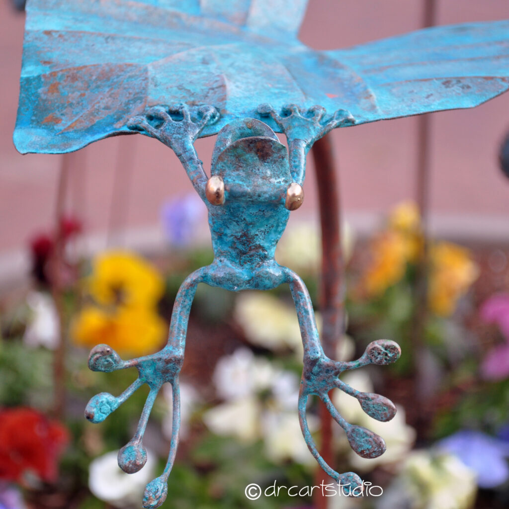 Photo of a hand made copper frog, trying to climb on a leaf.