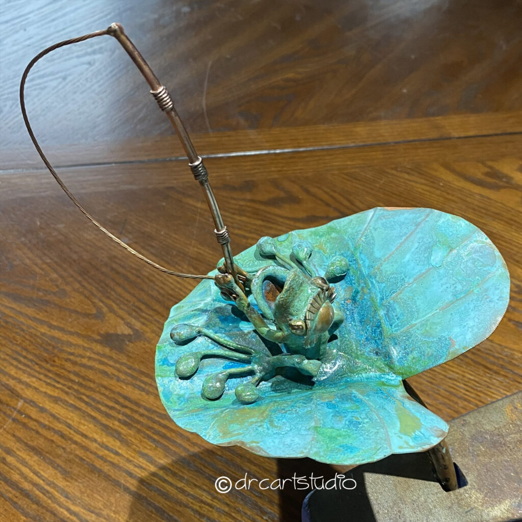 Photo of a hand made copper frog, holding a fishing rod and standing on a leaf.