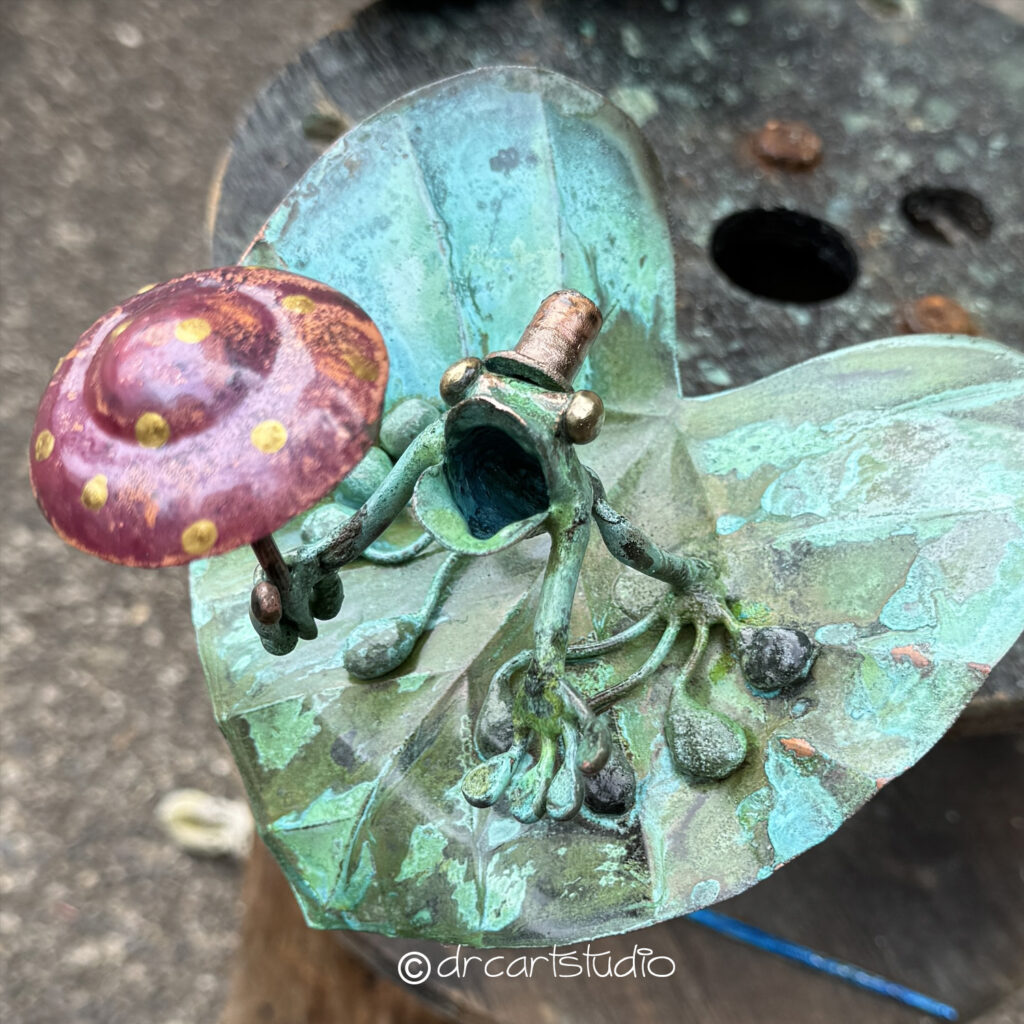 Photo of a hand made copper frog, holding an umbrella and standing on a leaf.